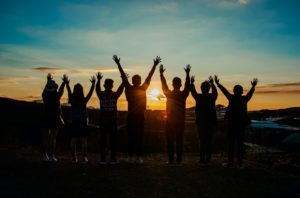 Eine Gruppe von Menschen blickt dem Sonnenuntergang entgegen und reißt die Arme in die Höhe.