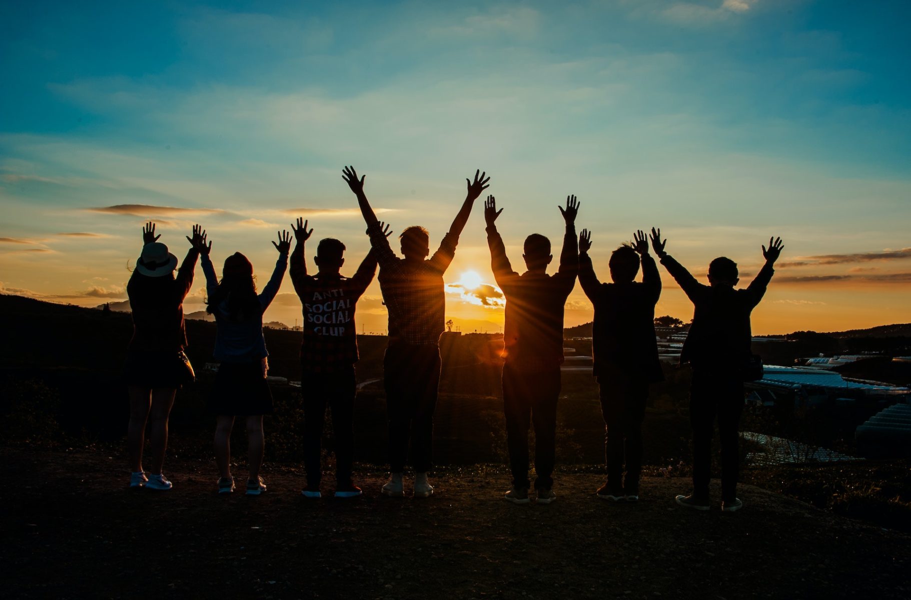 Eine Gruppe von Menschen blickt dem Sonnenuntergang entgegen und reißt die Arme in die Höhe.