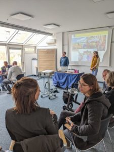 Die beiden Referenten stehen vor der Powerpoint Präsentation während die Teilnehmenden sich in kleinen Gruppen beraten